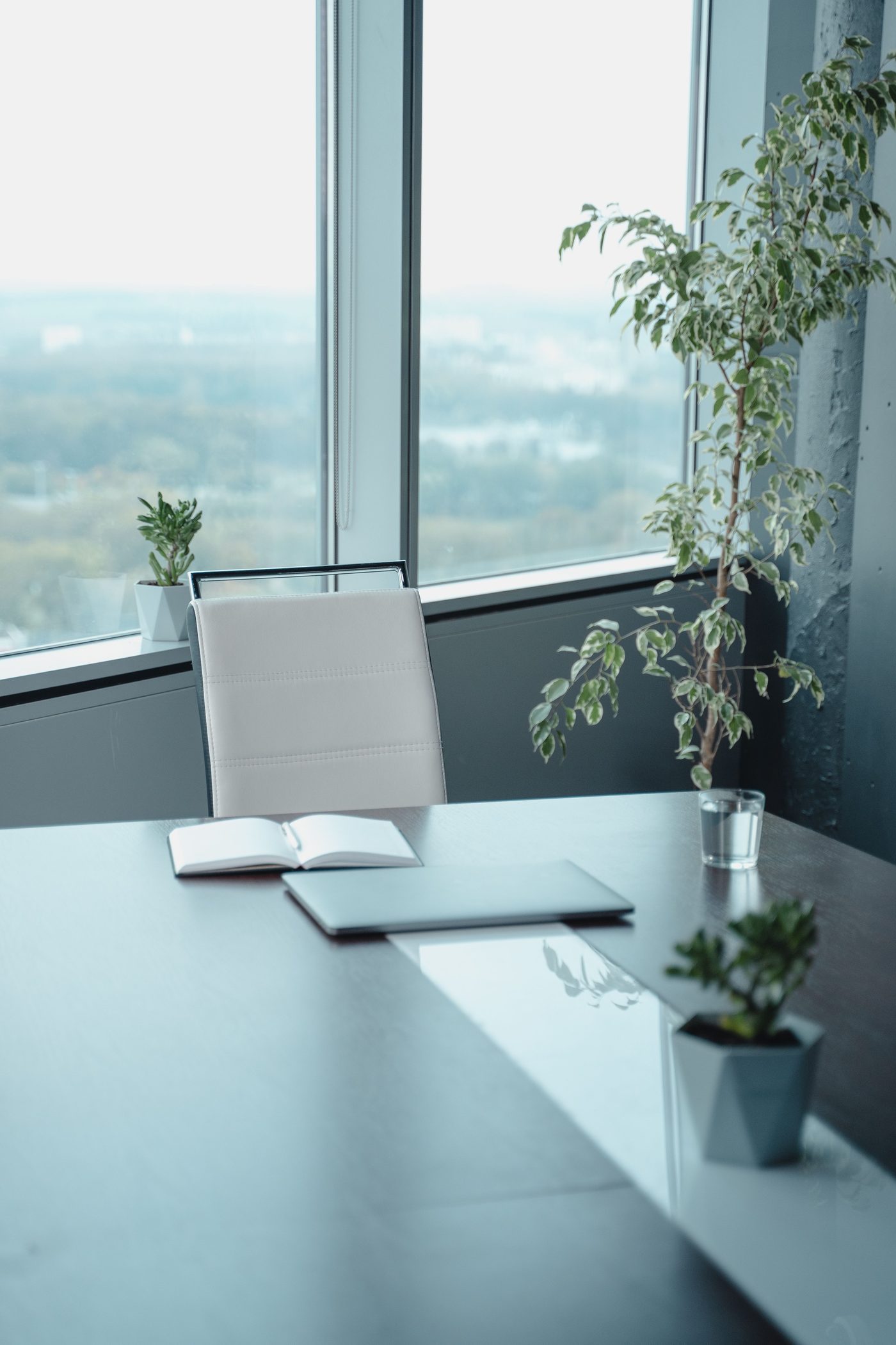 An uncluttered executive desk beside tall corner windows, with a notebook, laptop, and a soft daylight view.